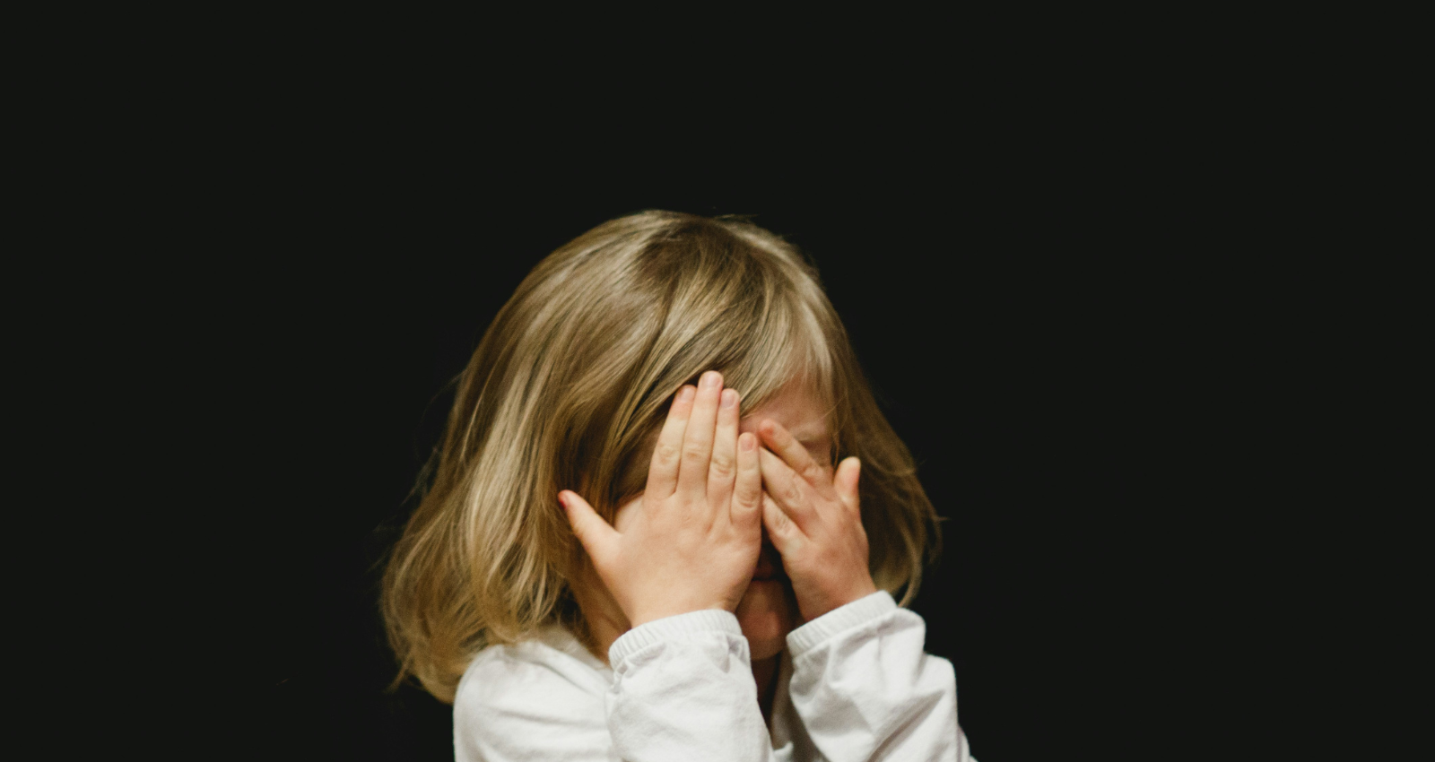 young girl with blonde hair holding her hands over her face. Background is black.