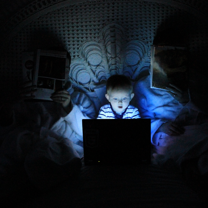 Internet Safety. A baby sitting in between parents in bed, the room is pitch black. A light is shining on his face from looking at a computer screen.
