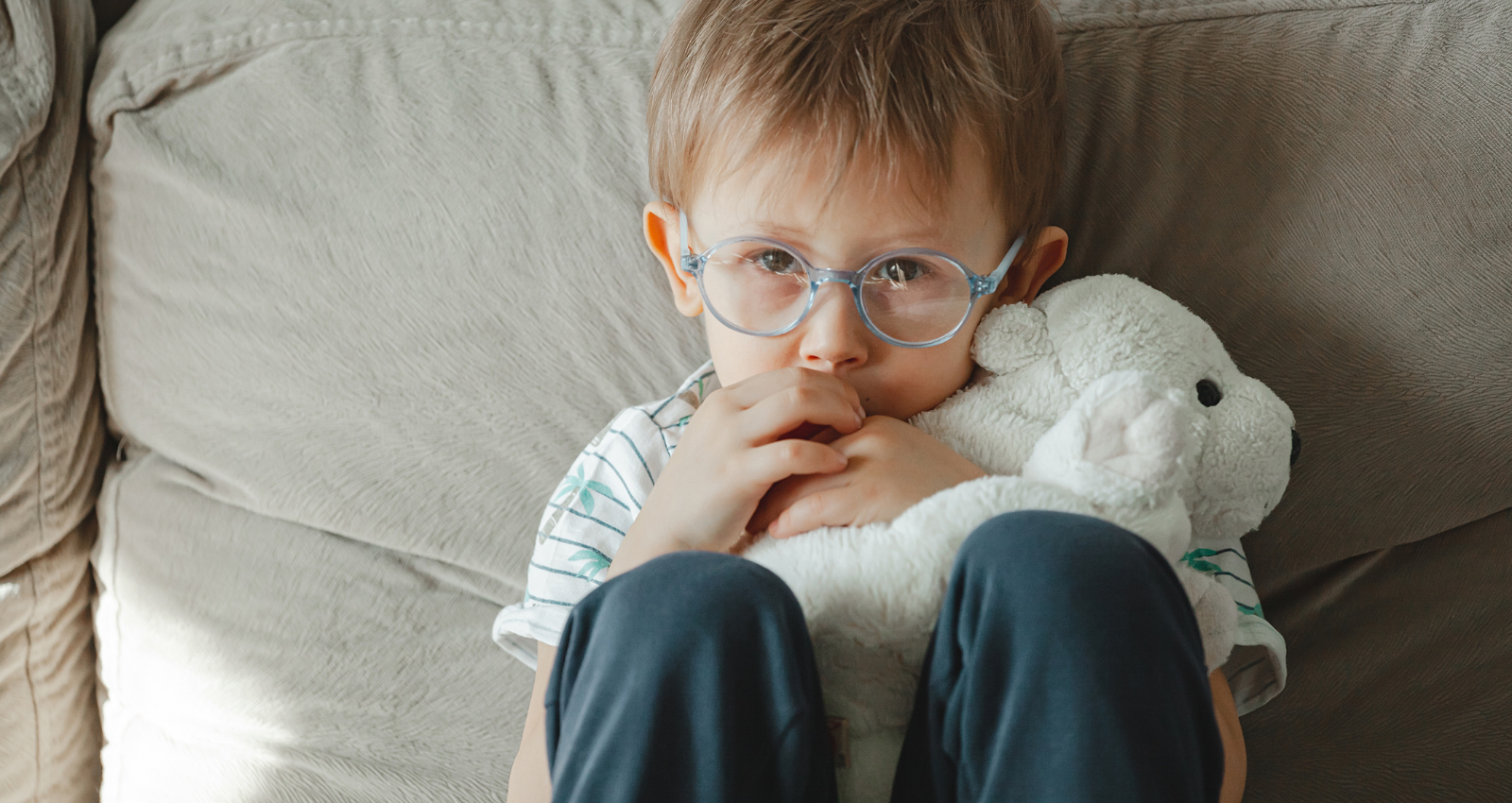 young boy sitting on sofa with knees up by chest, hands in mouth looking sad. Comforted by a white plush toy