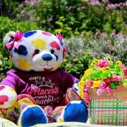 Photo of a colourful teddy bear with a gift box all wrapped up. Sitting in a summer garden setting with bushes behind.